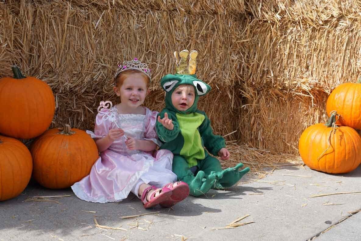 A little girl dressed as a princess and a little boy dressed as a frog are sitting next to pumpkins.