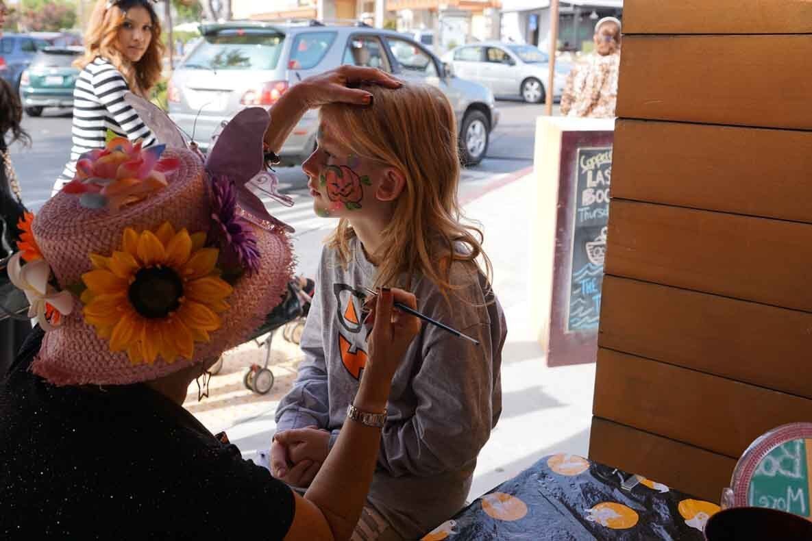 A little girl is getting her face painted by a woman wearing a hat with a sunflower on it.