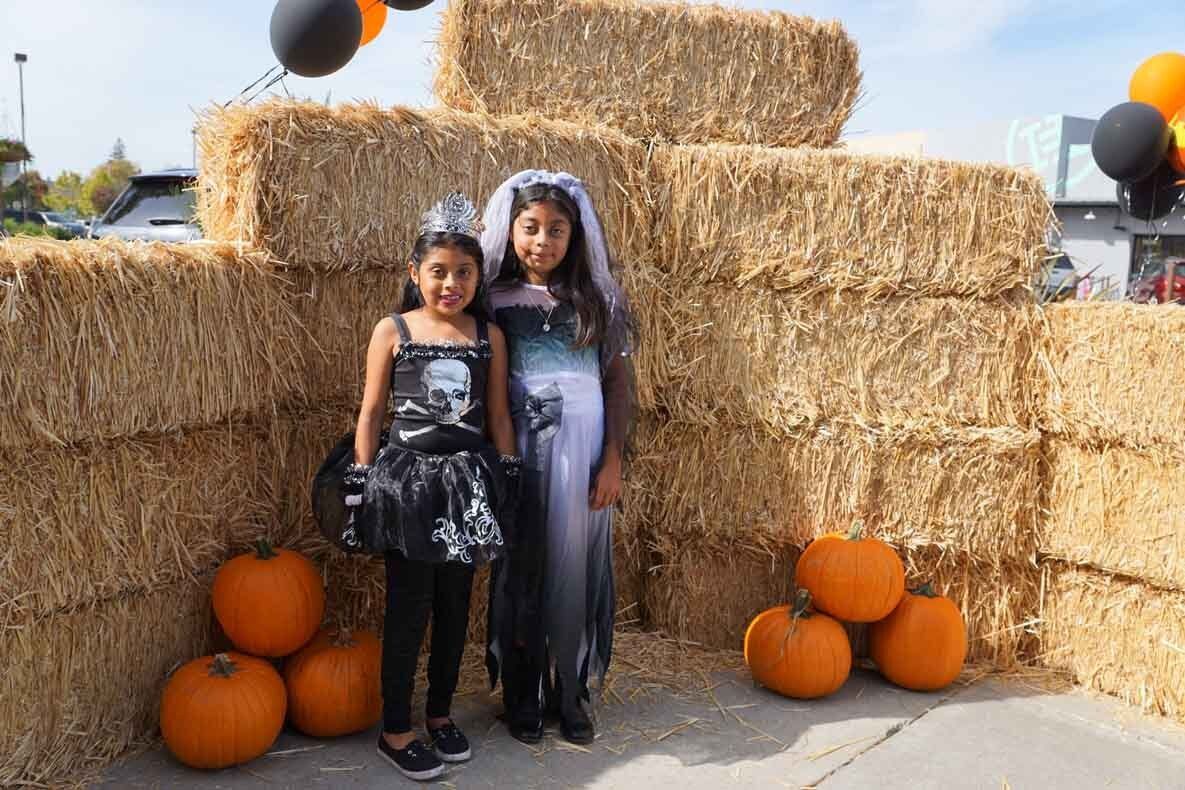Two little girls in halloween costumes are standing next to hay bales and pumpkins.