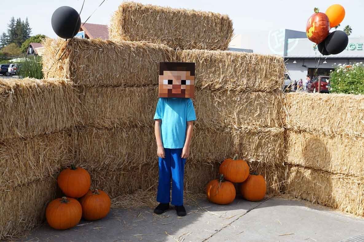 A boy wearing a minecraft mask is standing in front of hay bales and pumpkins.