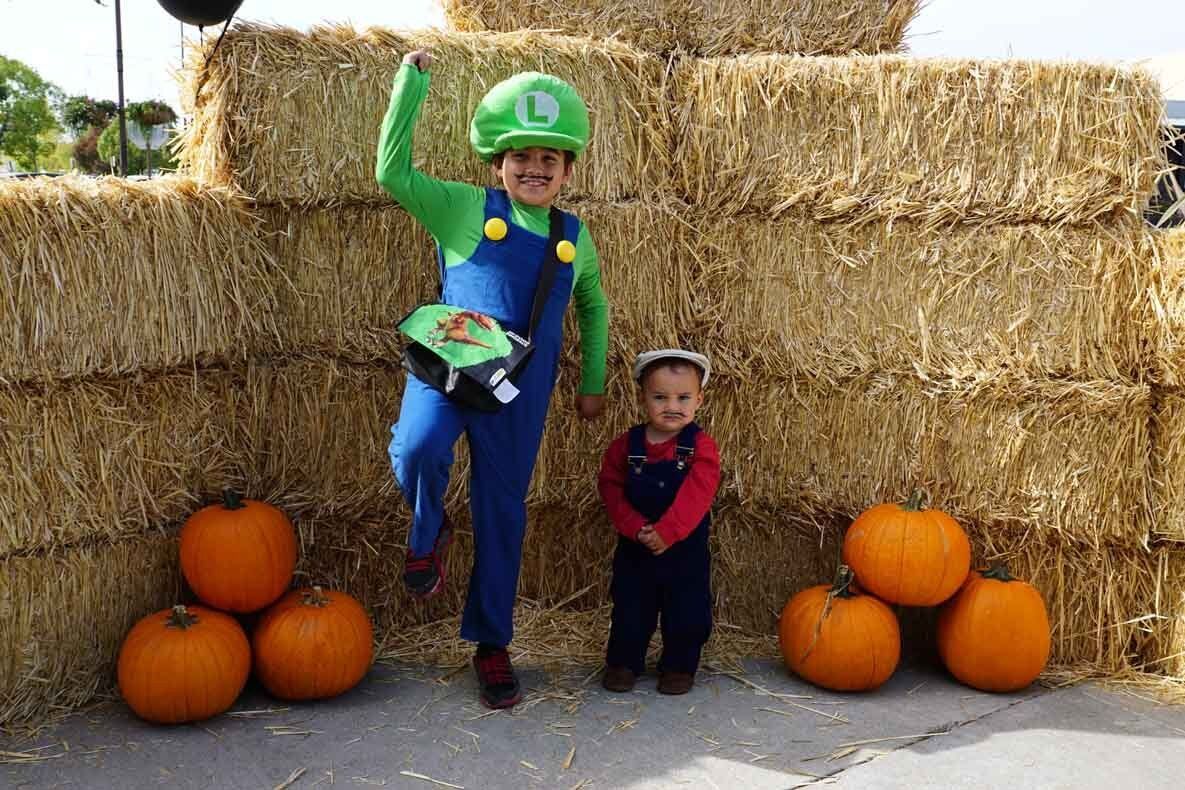 Two children dressed in costumes are standing in front of hay bales and pumpkins.