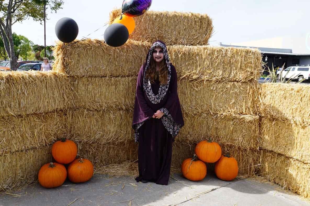 A woman in a witch costume is standing next to hay bales and pumpkins.