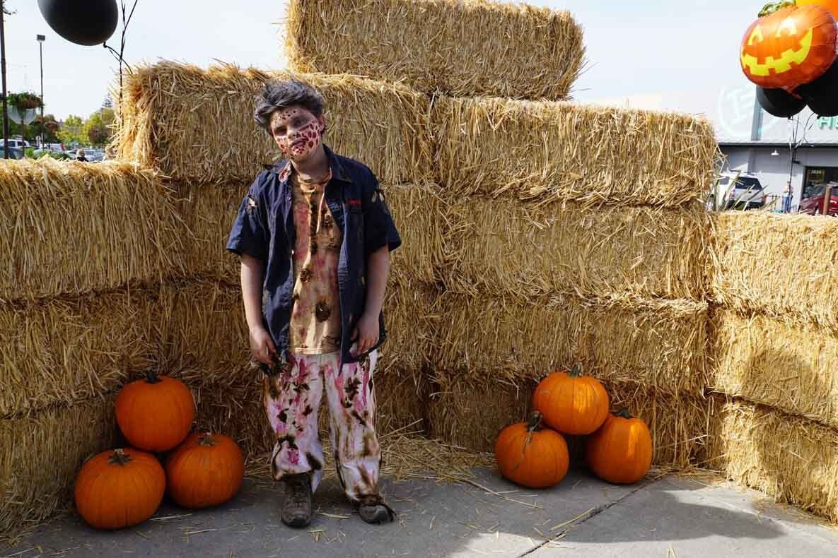 A boy in a zombie costume is standing in front of hay bales and pumpkins.