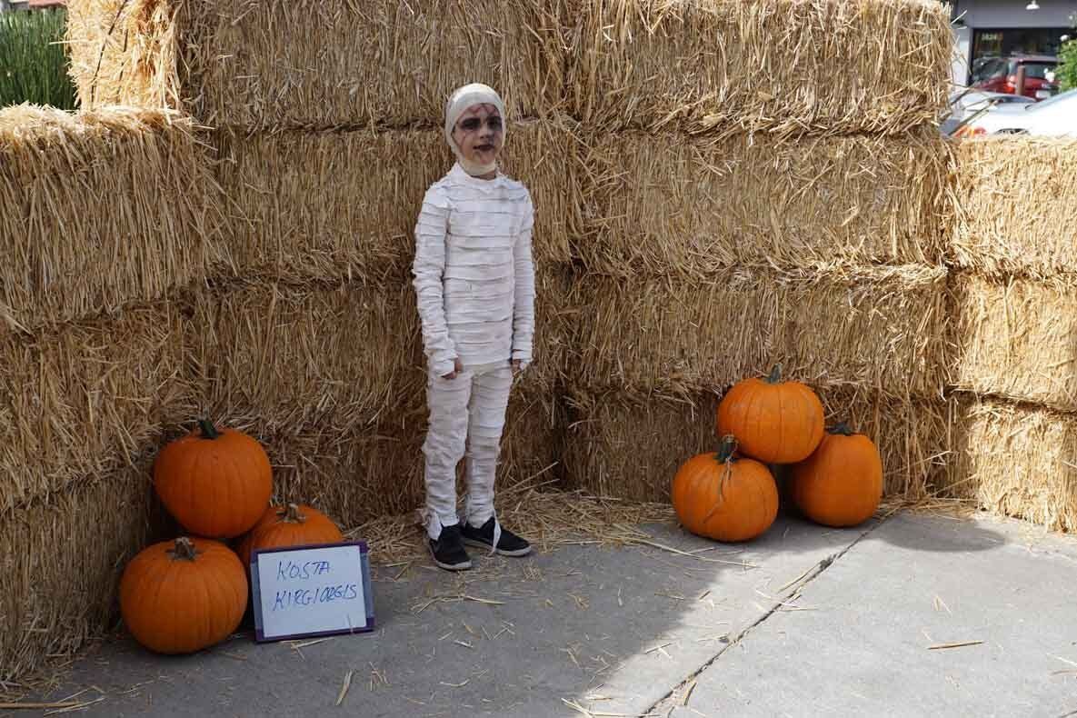 A child dressed as a mummy is standing next to hay bales and pumpkins.