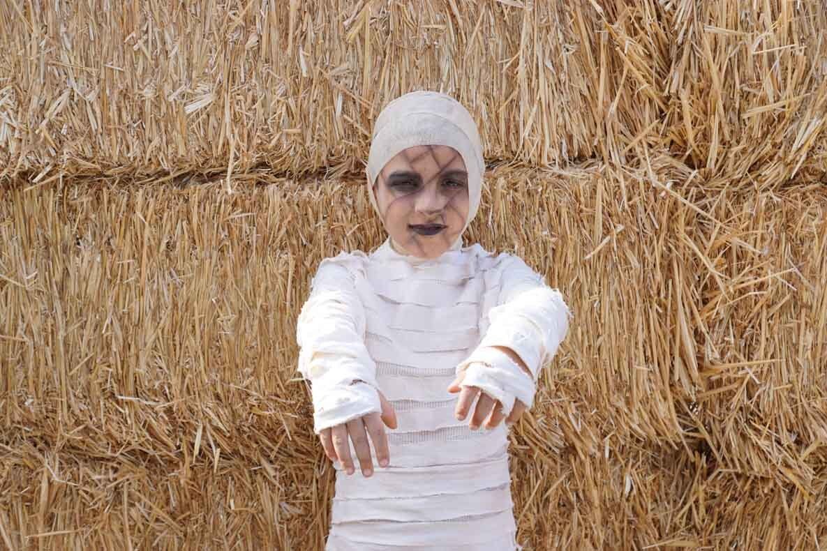 A child dressed as a mummy is standing in front of a hay bale.
