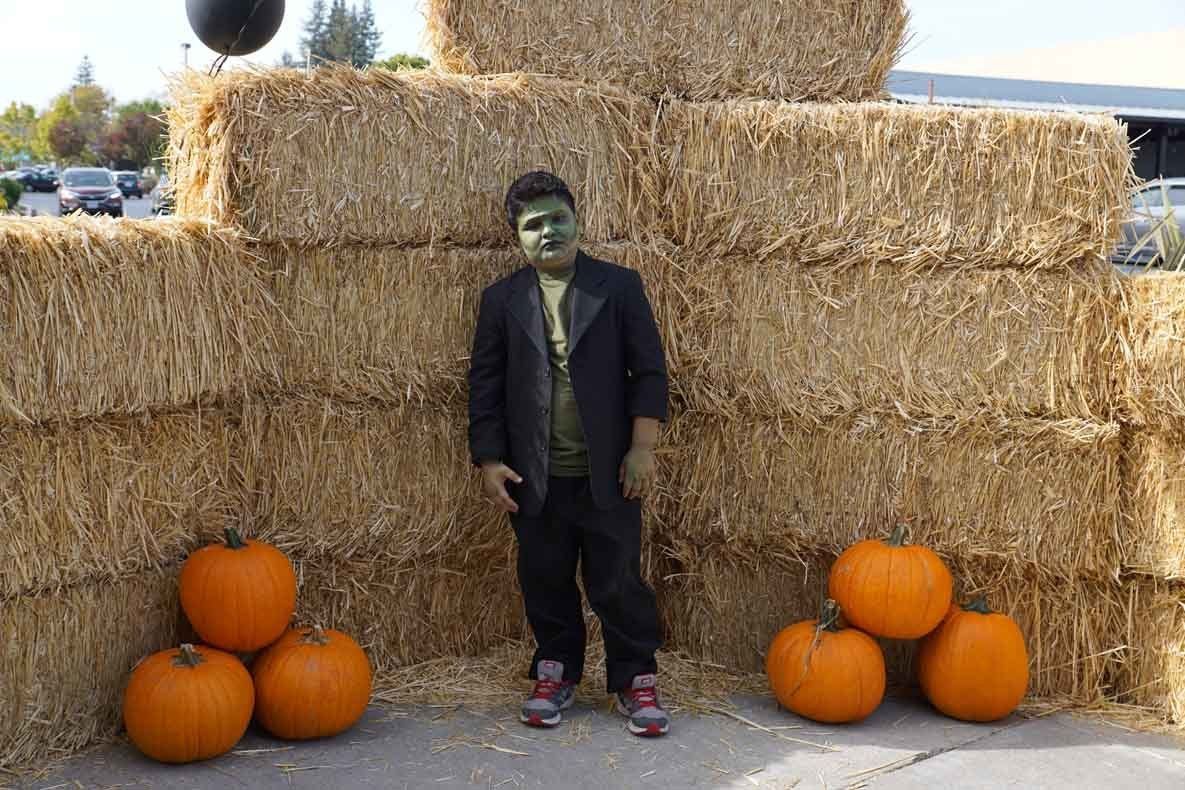 A boy in a frankenstein costume is standing next to a pile of hay bales and pumpkins.