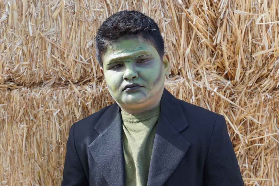 A young boy with green face paint is standing in front of a hay bale.
