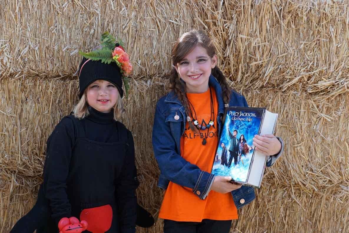 Two young girls in costumes are standing next to each other in front of a hay bale.