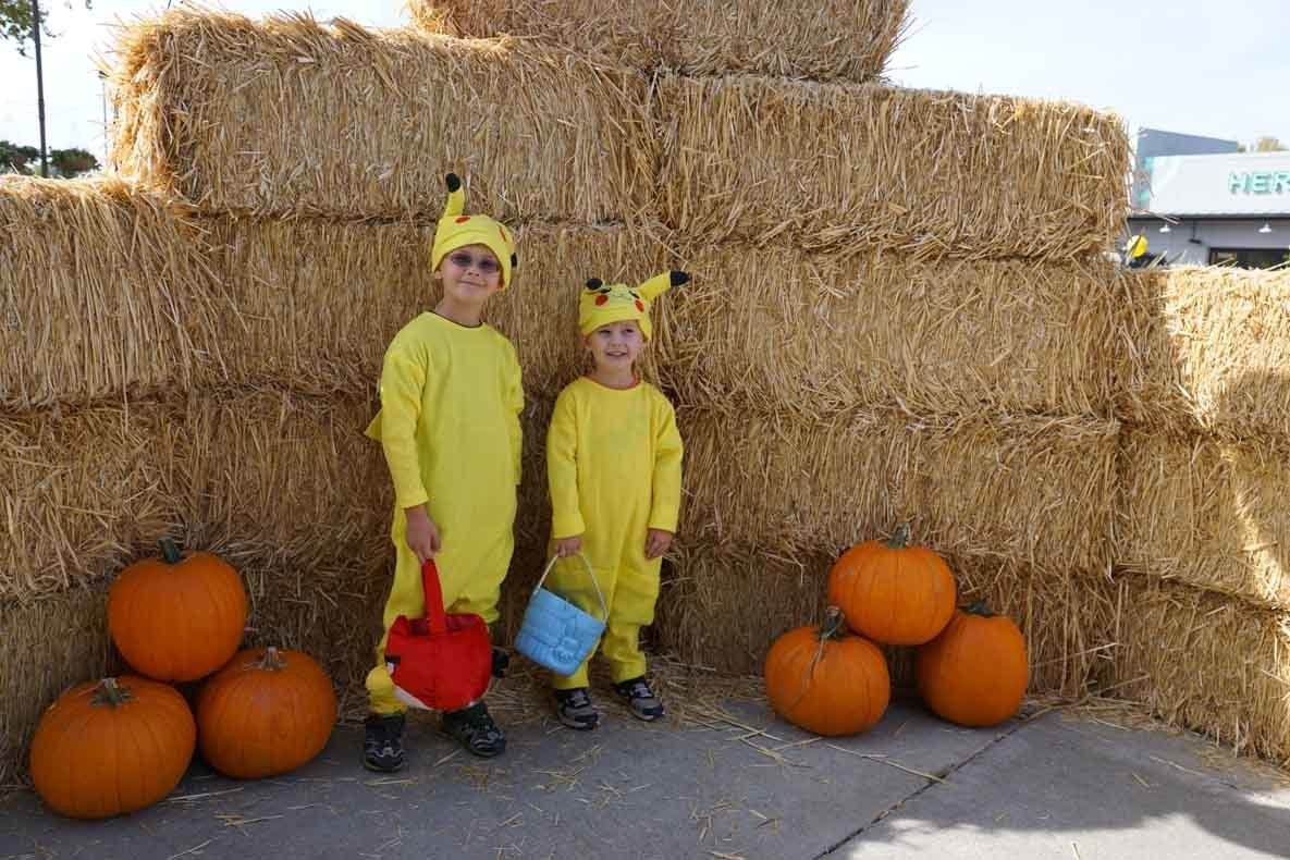 Two children in pikachu costumes are standing next to hay bales and pumpkins.