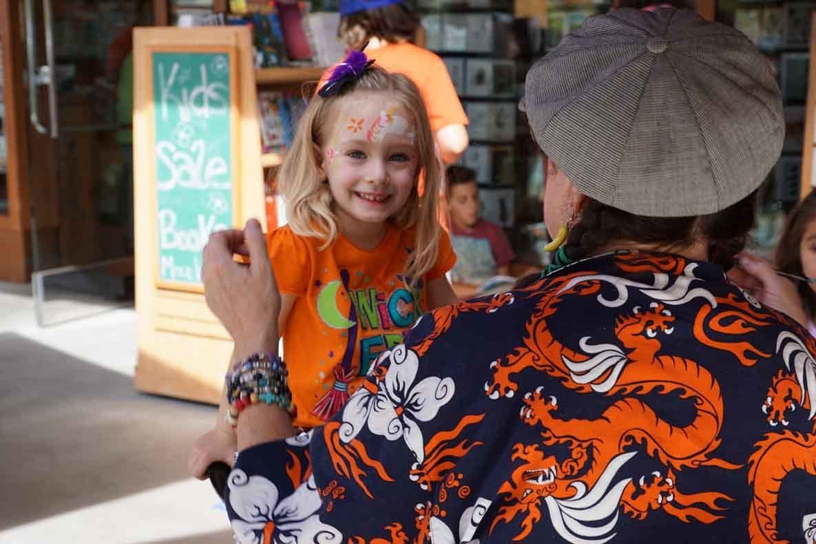 A man is painting a little girl 's face in front of a sign that says kids sale