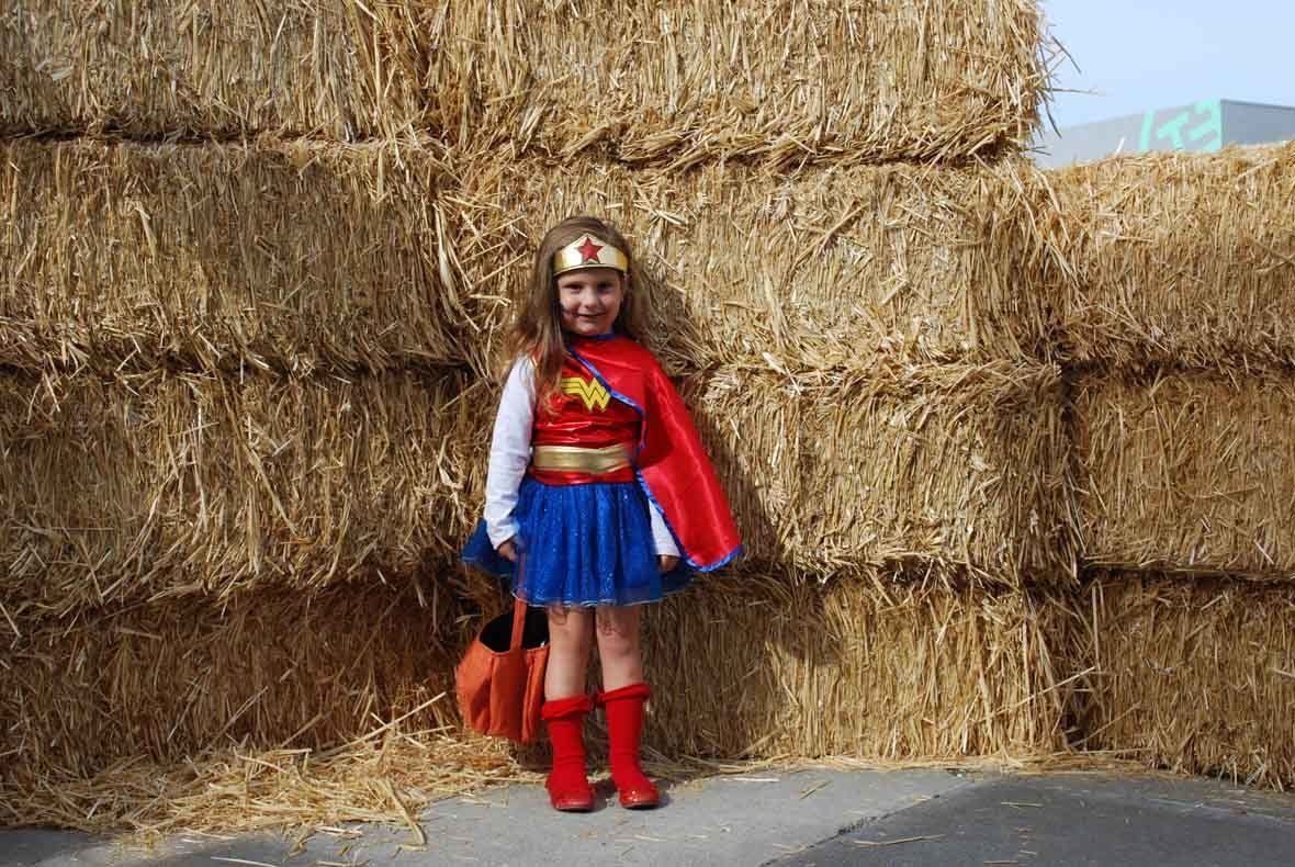 A little girl in a wonder woman costume is standing in front of a hay bale.