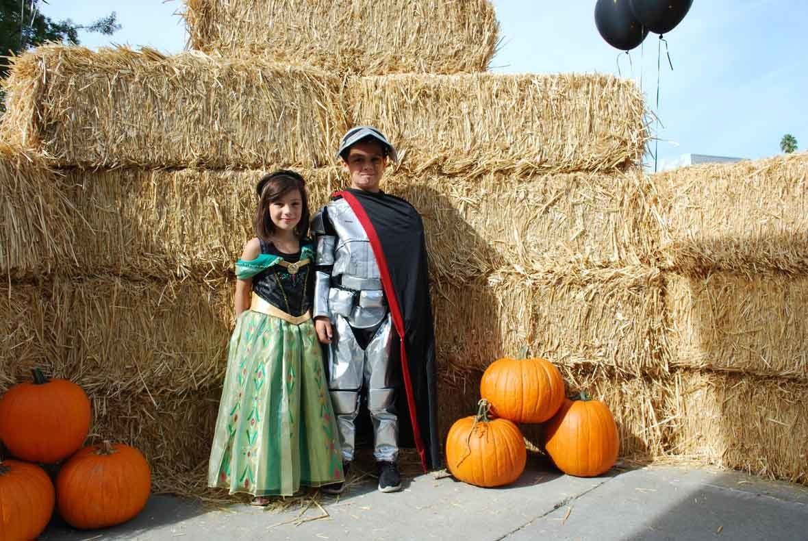 A boy and a girl in costumes are standing in front of hay bales and pumpkins.