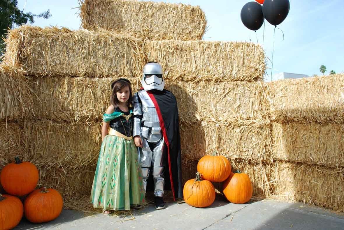 Two children in costumes are standing in front of hay bales and pumpkins.