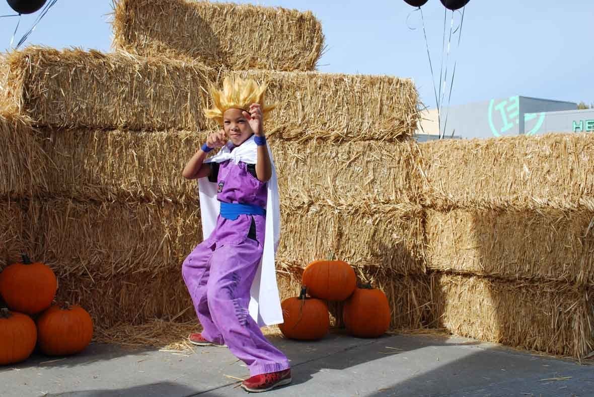 A person in a costume is standing in front of hay bales and pumpkins.