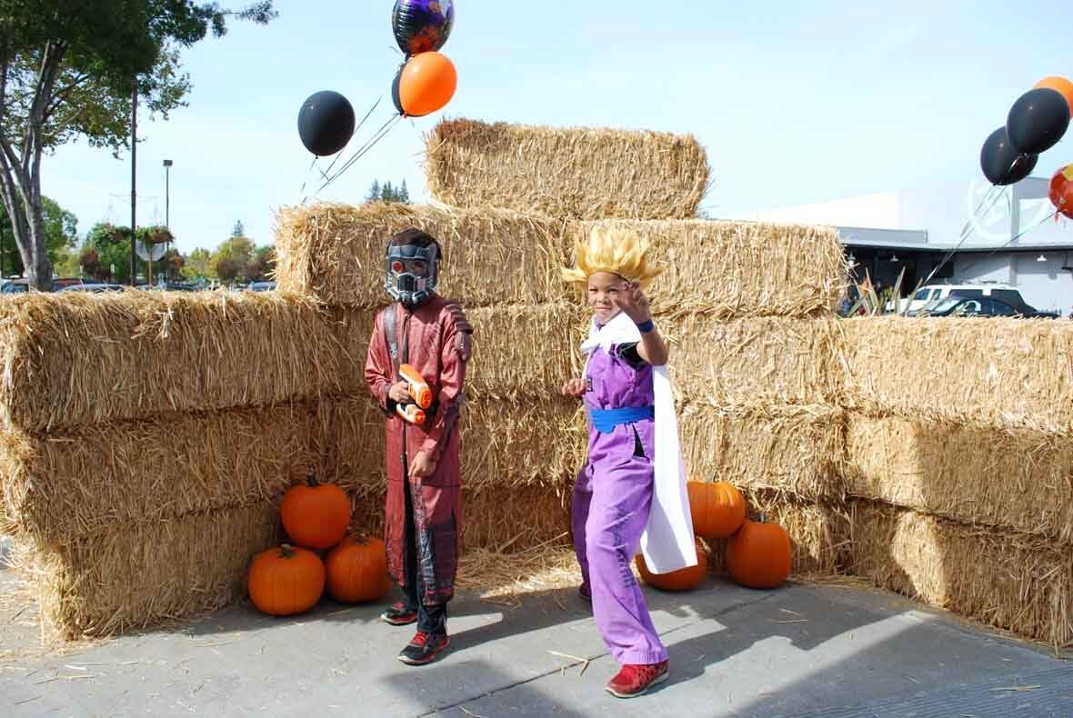 Two people in costumes are standing in front of hay bales and pumpkins.