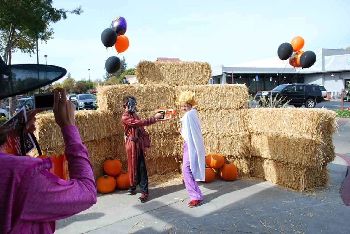 A man is taking a picture of a zombie and a woman in front of hay bales and pumpkins.