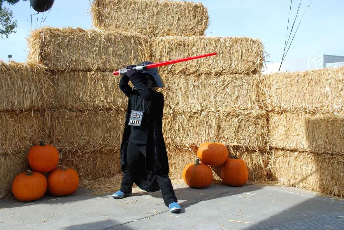 A child in a star wars costume is holding a lightsaber in front of hay bales and pumpkins.