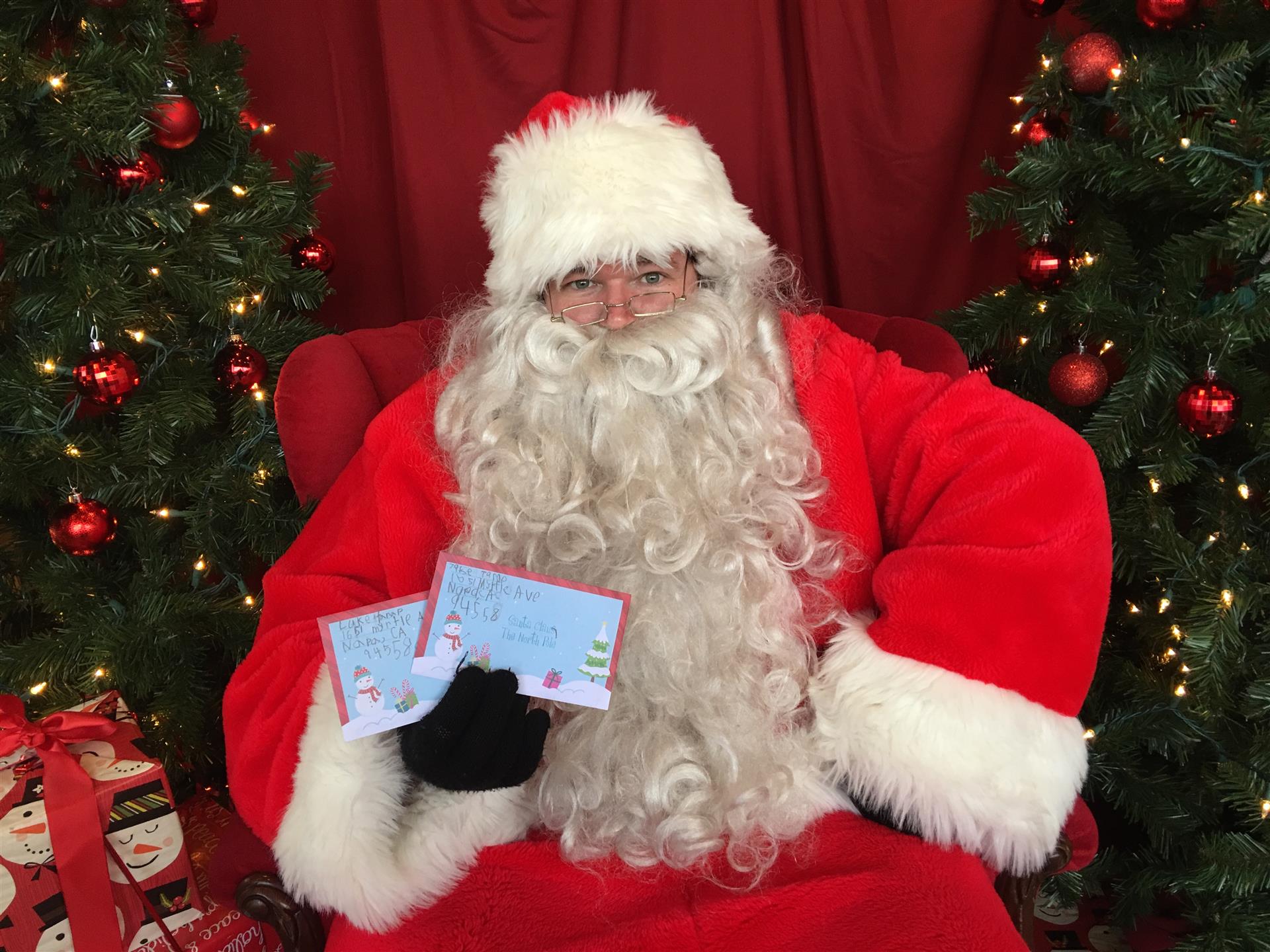 Santa claus is sitting in a chair holding two cards in front of a christmas tree.