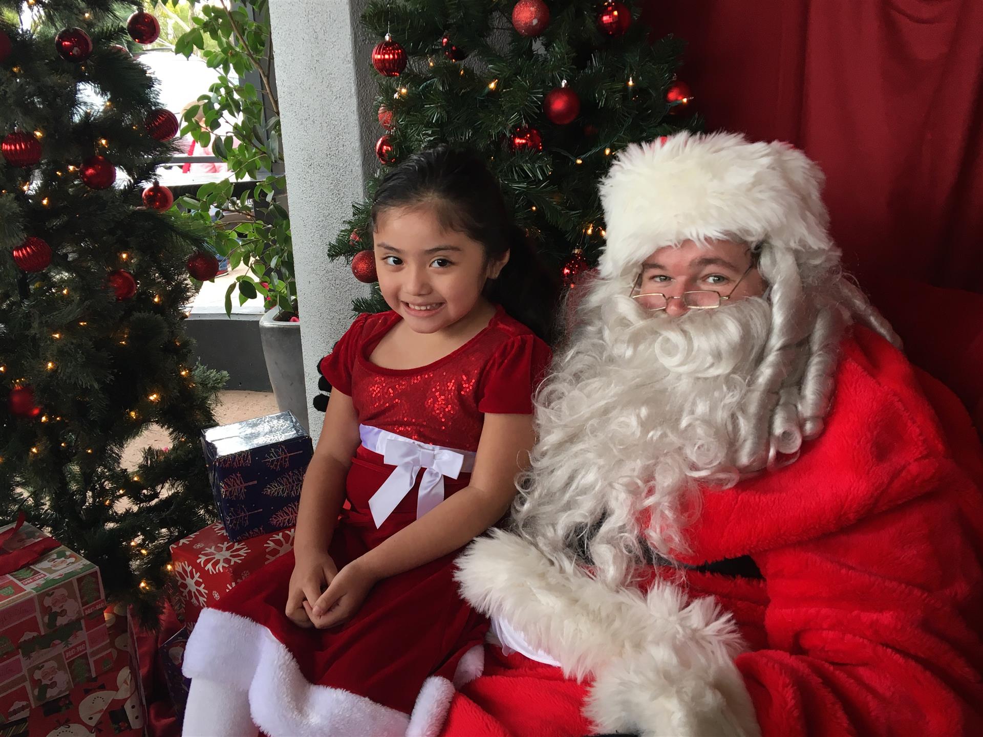 A little girl is sitting next to santa claus in front of a christmas tree.