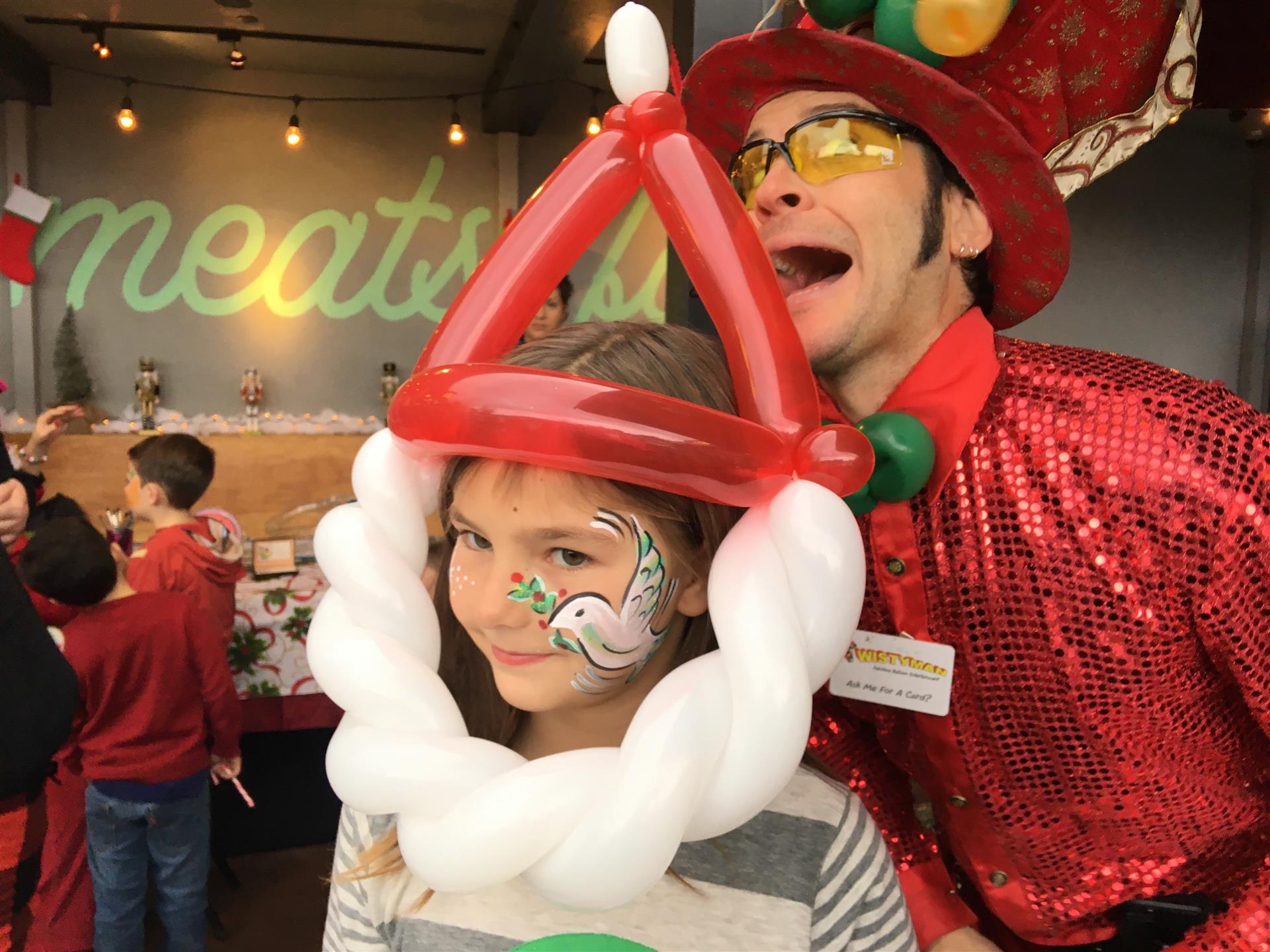 A man in a red hat is standing next to a little girl with balloons painted on her face.