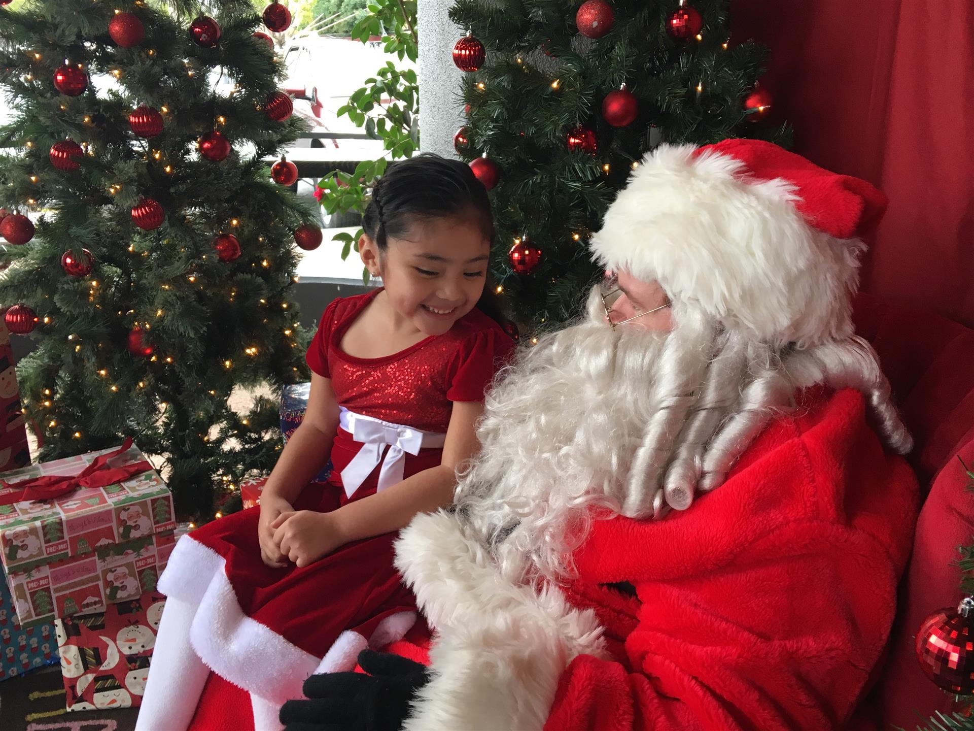 A little girl is sitting next to santa claus in front of a christmas tree.