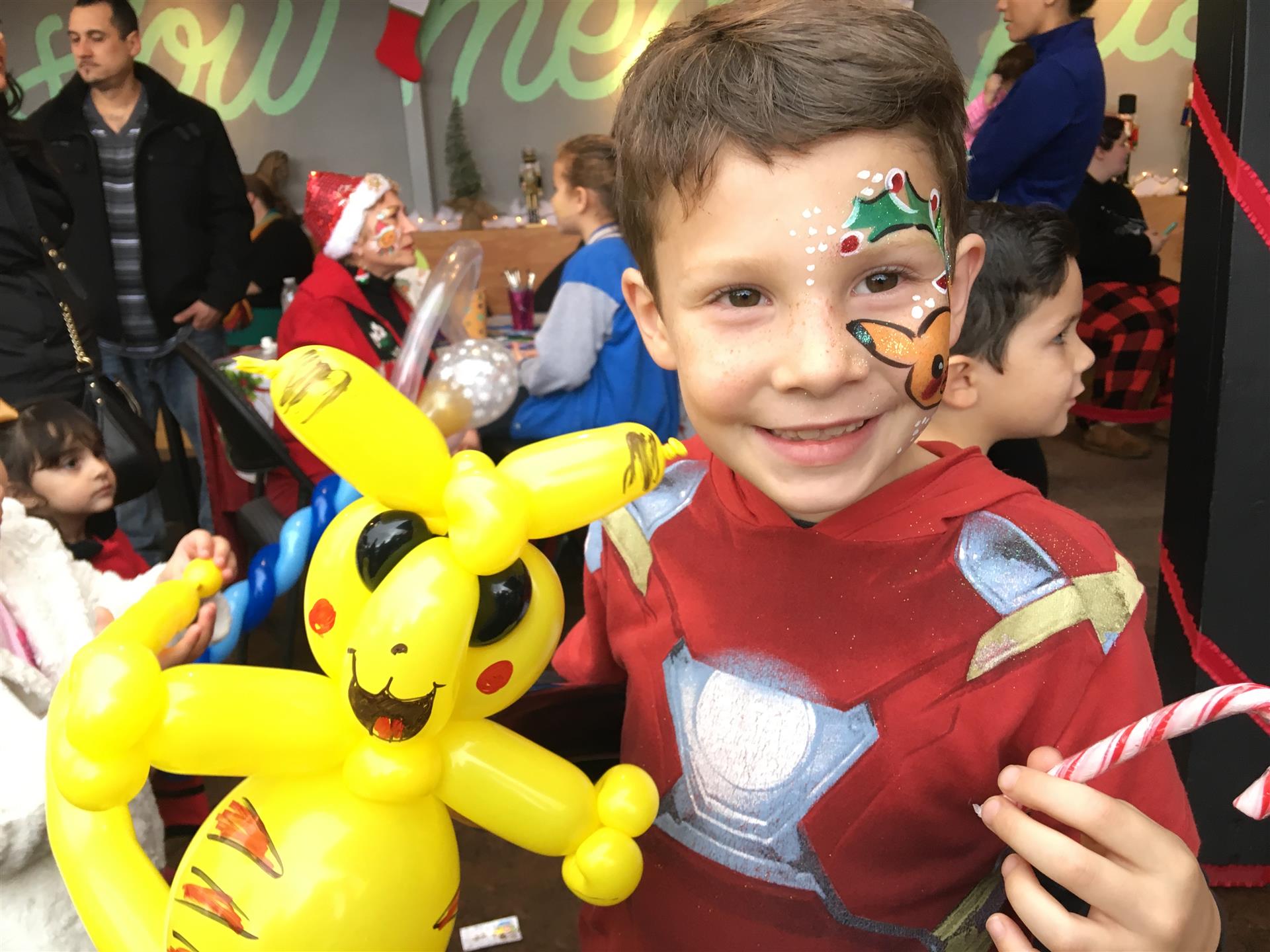 A young boy with balloons painted on his face is holding candy canes and a pikachu balloon.