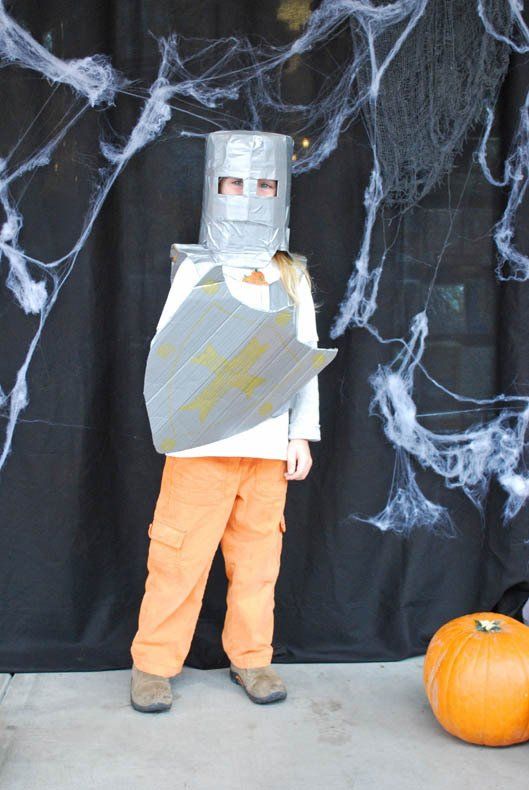 A child in a cardboard knight costume is standing next to a pumpkin.