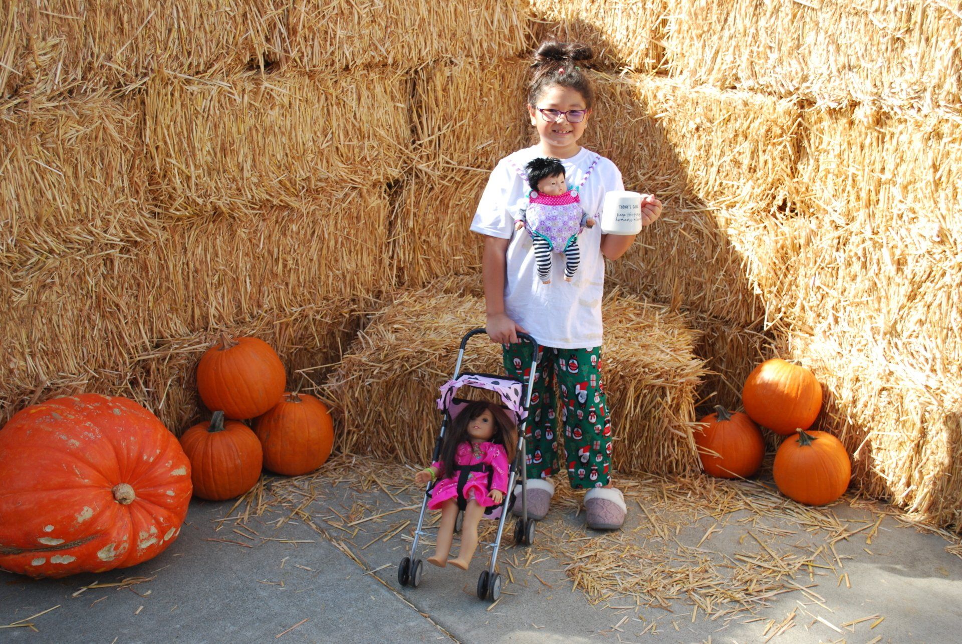 A little girl is holding a doll in a stroller in front of hay bales and pumpkins.