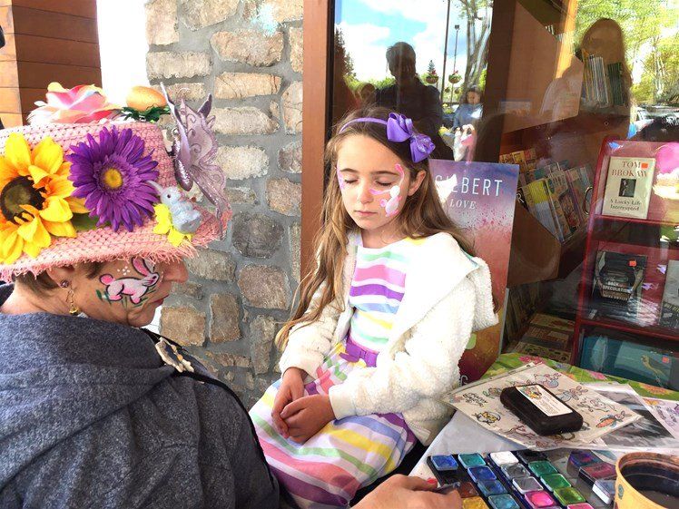 A little girl is getting her face painted by a person wearing a hat with flowers on it