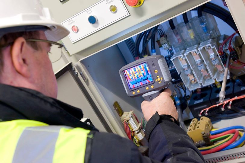 Un trabajador con casco y chaleco reflectante utiliza una cámara termográfica infrarroja en un panel eléctrico.