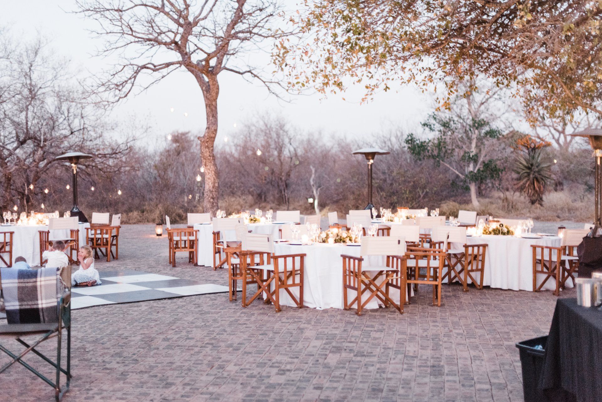 Outdoor event setup with white-clothed tables and wooden chairs under trees at dusk