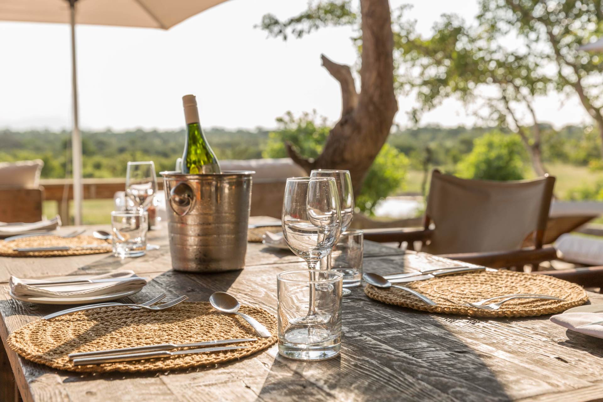 Outdoor dining table set with woven placemats, wine glasses, champagne bucket, and umbrella in a sunny garden