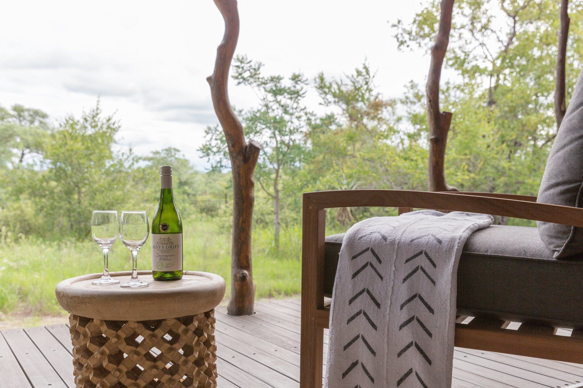 Patio with wine bottle and glasses on a small table beside a gray chair and blanket overlooking greenery