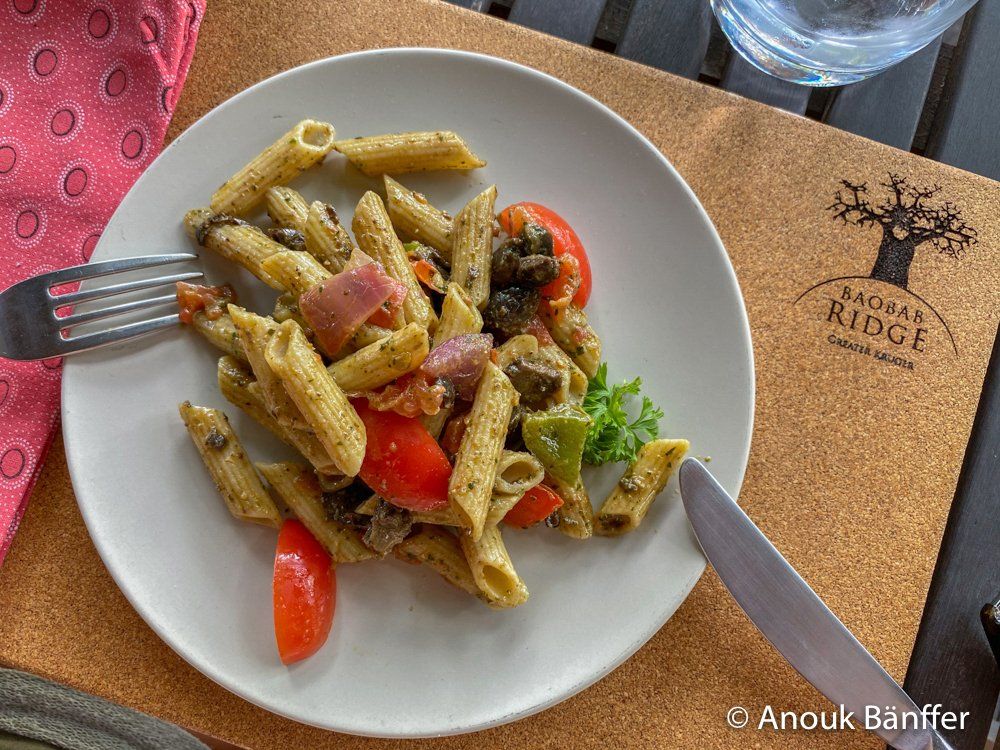 Pesto penne with cherry tomatoes on a white plate at a café table, with a fork and napkin nearby