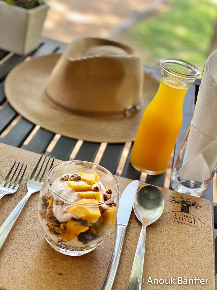 Outdoor café table with fruit dessert, orange drink, spoon, fork, and a straw hat in the background