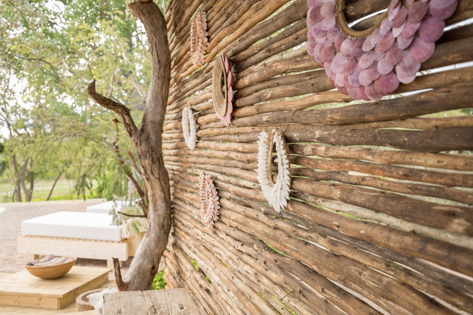 Rustic woven wall with hanging decorative circles beside a shaded outdoor seating area under trees