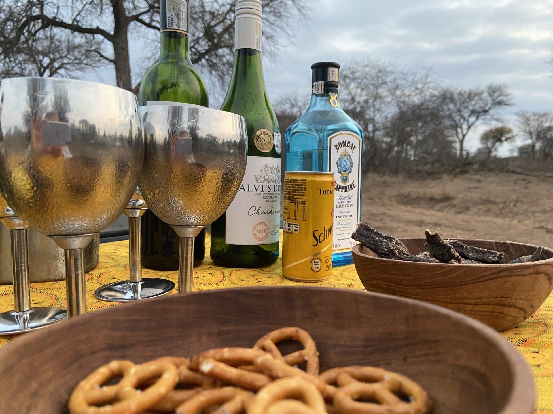 Outdoor table with wine bottles, glasses, a blue liqueur bottle, snacks, and a cloudy sky backdrop