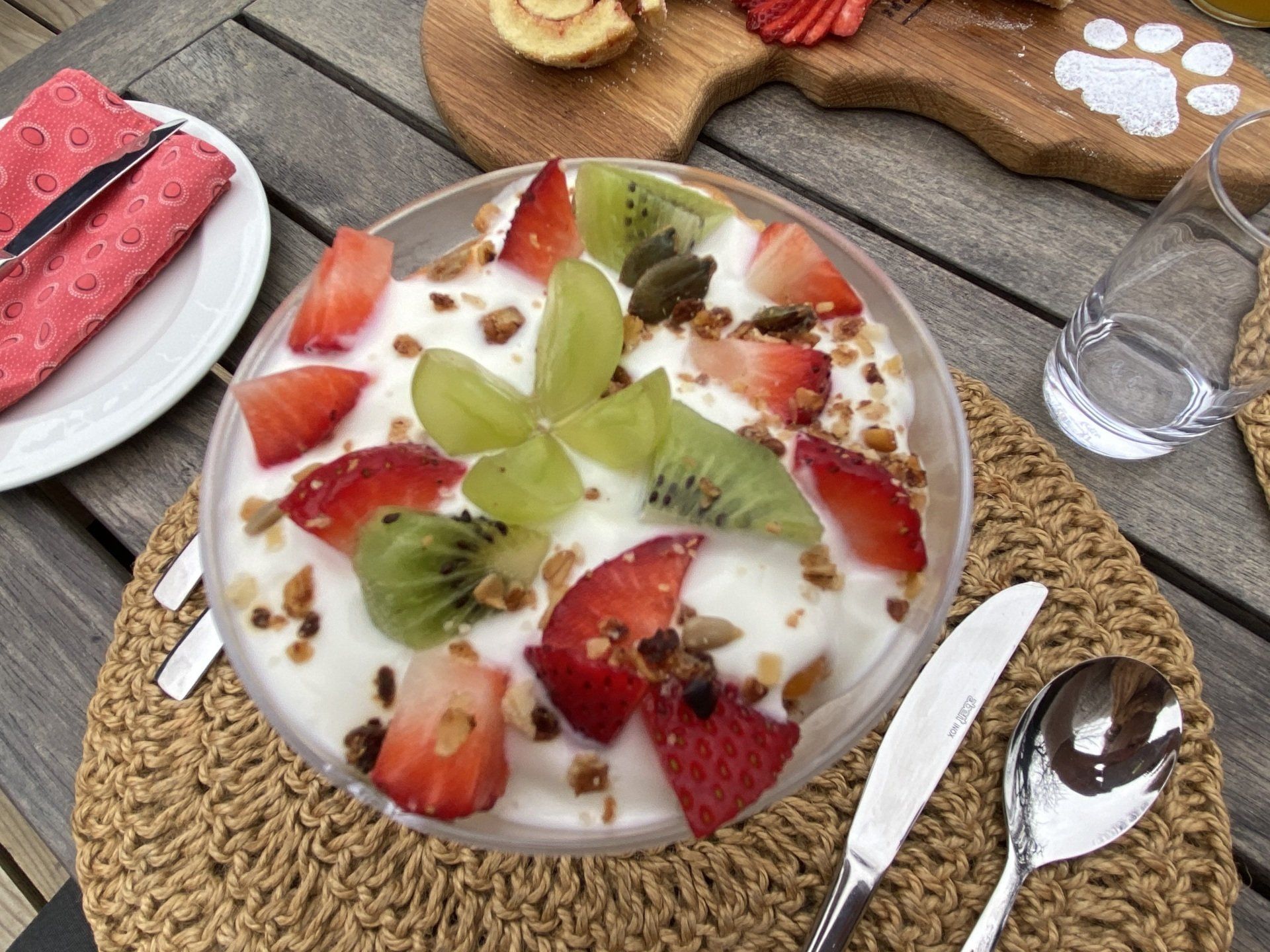 Fruit-topped yogurt dessert on a table with spoon, knife, and glasses nearby