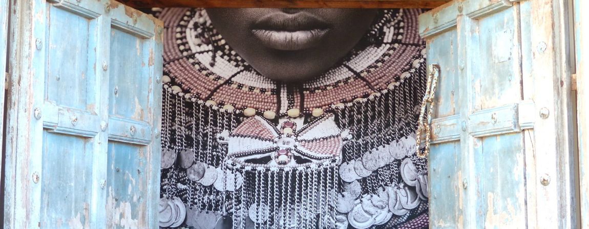 Close-up of a person’s face framed by weathered blue wooden doors, wearing a patterned beaded necklace.