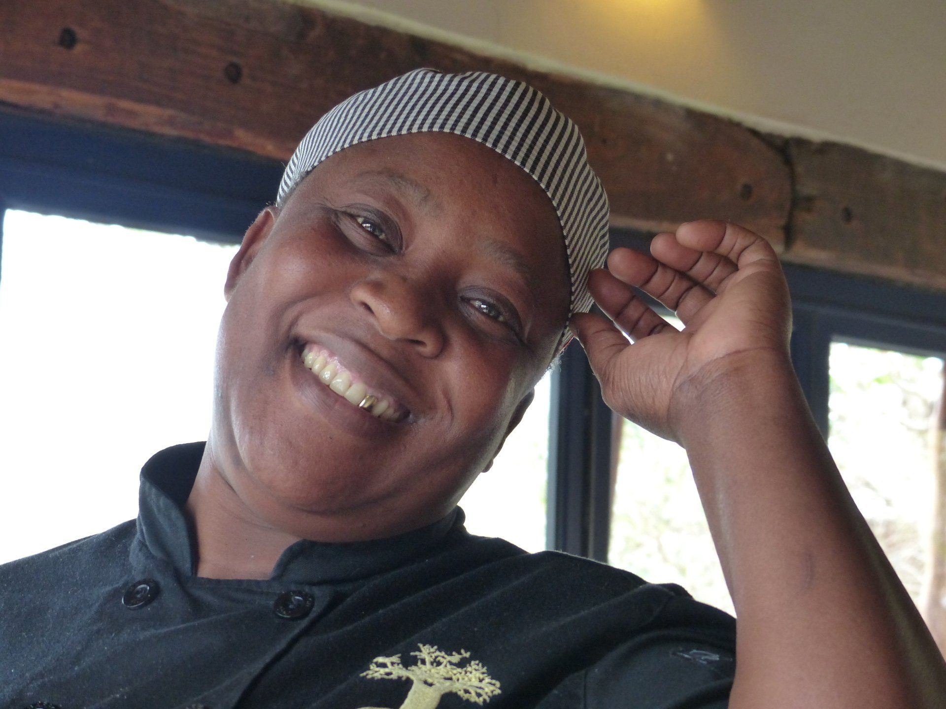 Smiling person in a black shirt and striped headscarf, leaning near a window inside a wooden room