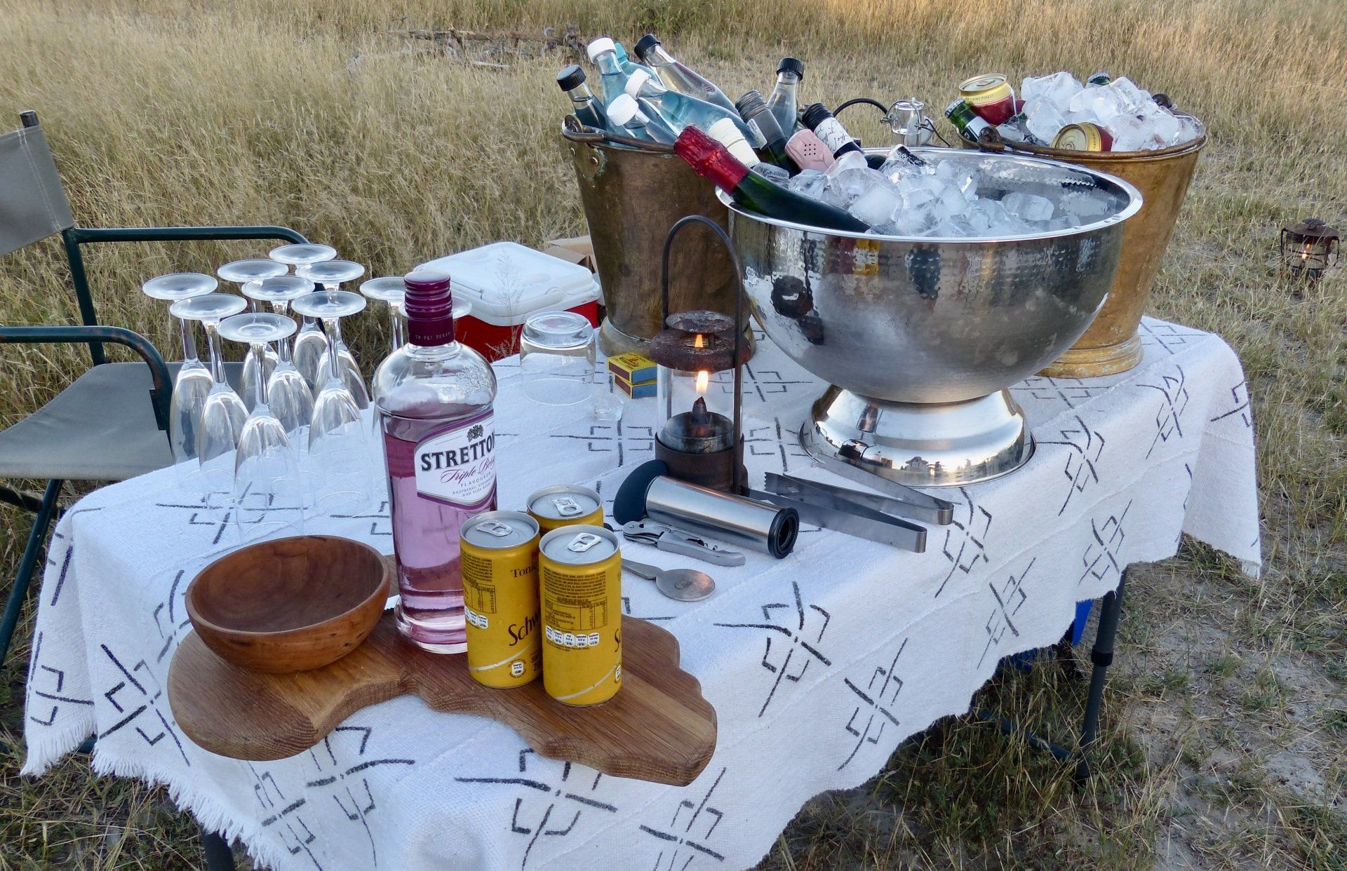 Outdoor drink station with bottles, cans, bowls, and an ice-filled tub on a table with a patterned cloth