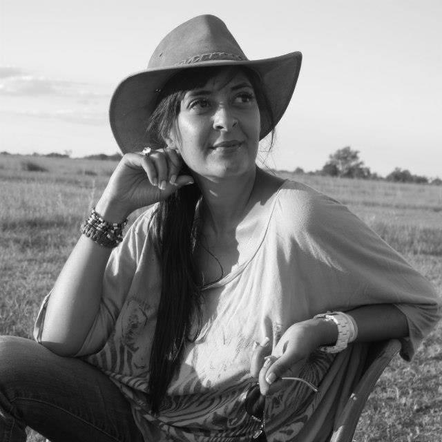 Woman in a cowboy hat sitting outdoors, smiling with hand on chin in a field.