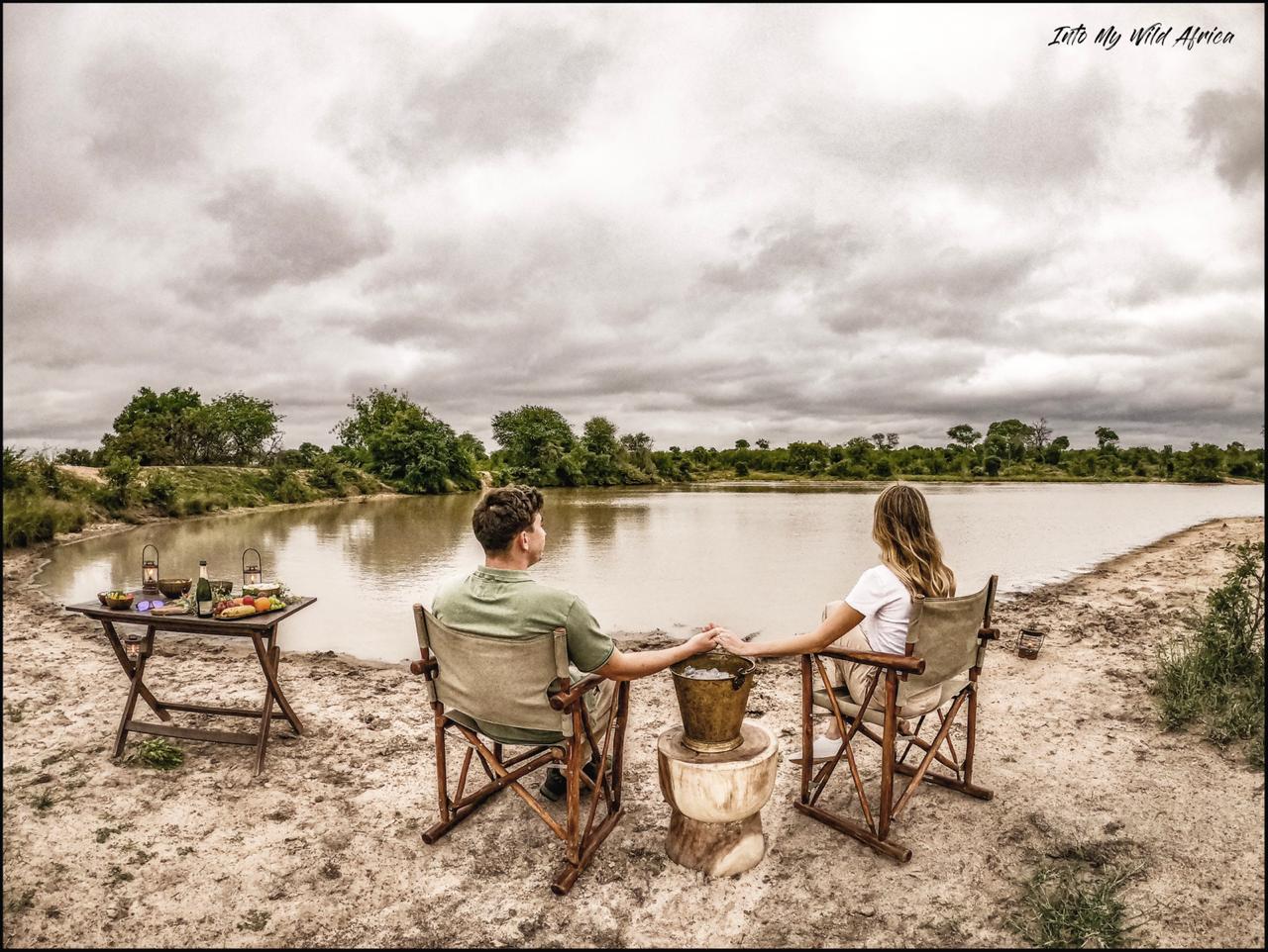 Two people sit by a flooded landscape under cloudy skies, with a small table and drinks nearby.