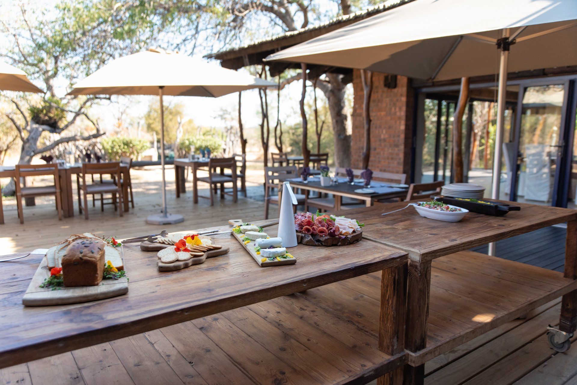 Outdoor wooden deck dining area with long table set for a meal under umbrellas beside a lodge.