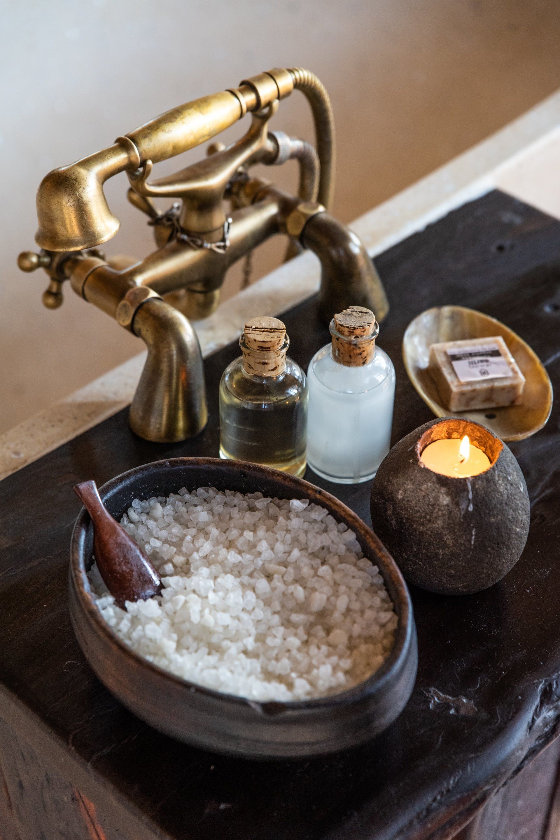 Spa setup with a brass faucet, bowl of bath salts, small bottles, and a lit candle on a dark tray