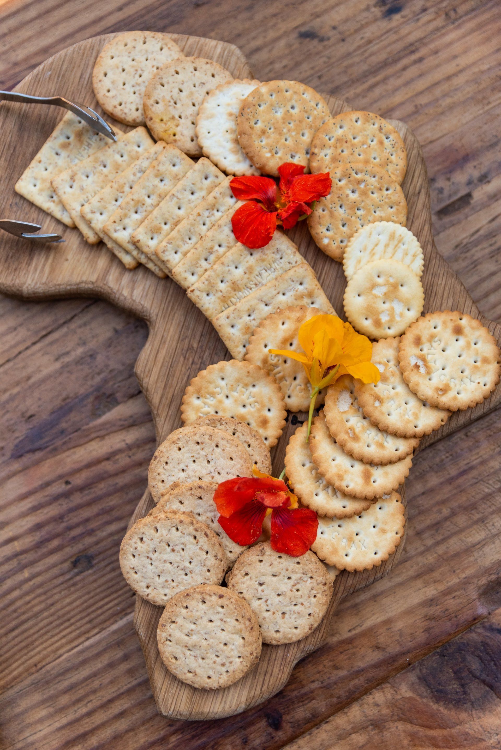 Cheese and crackers arranged on a wooden board with red and orange flowers as garnish