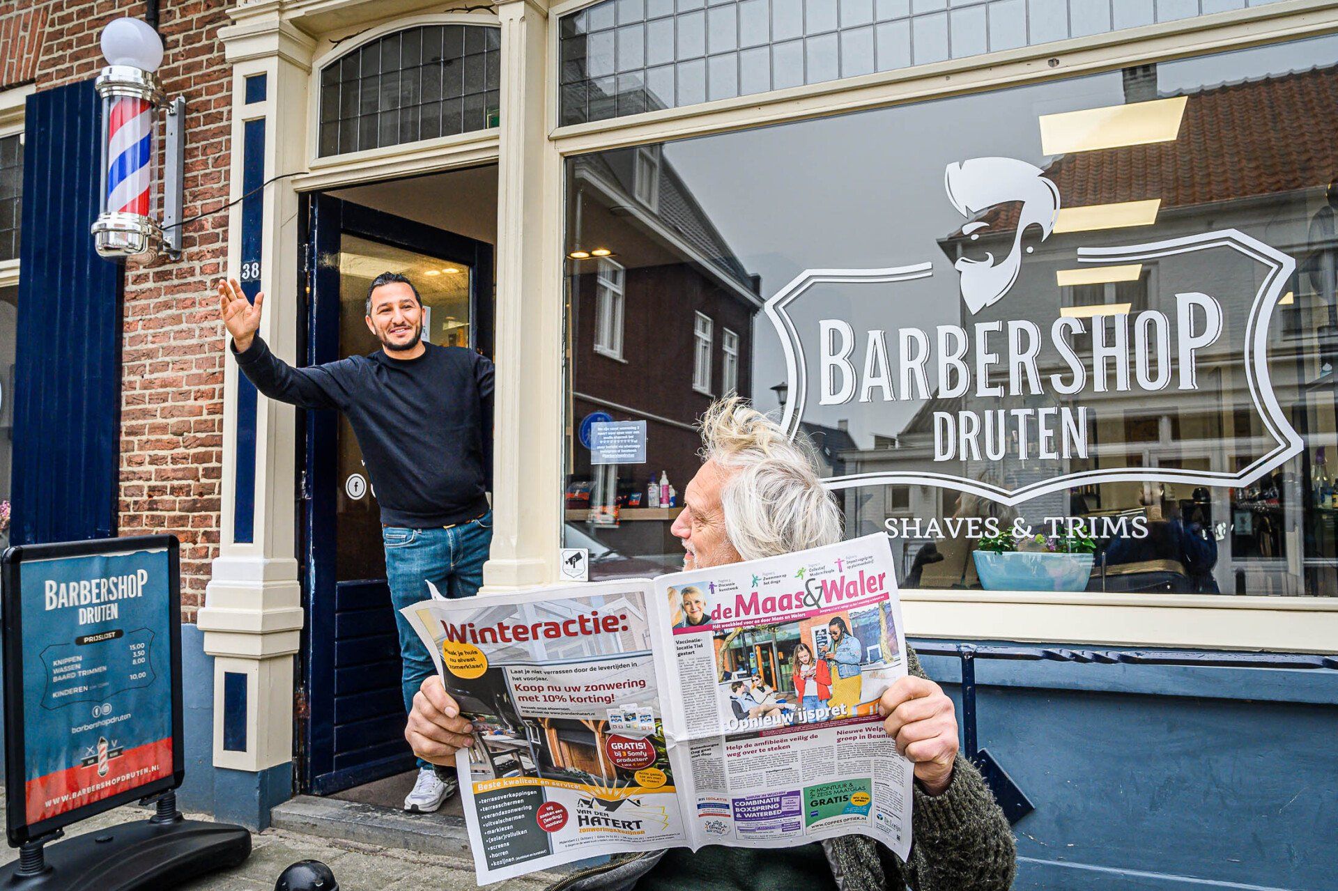 Een man staat voor een kapperszaak met een krant in zijn handen.
