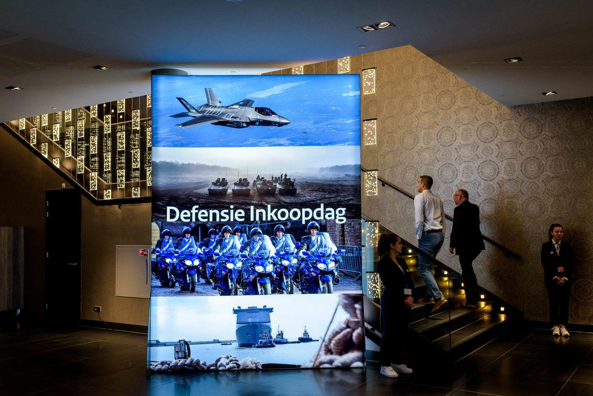 Mensen lopen de trap af voor een bord met de tekst 'Defensie Inkoopdag'