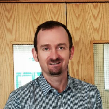 Smiling man in a blue checkered shirt standing in front of wooden doors.