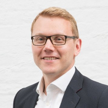 Portrait of a smiling person wearing glasses and a dark blazer against a white background